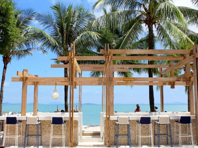 Otwarte bar na plaży z drewnianą konstrukcją, stołkami barowymi i palmami przy morzu.