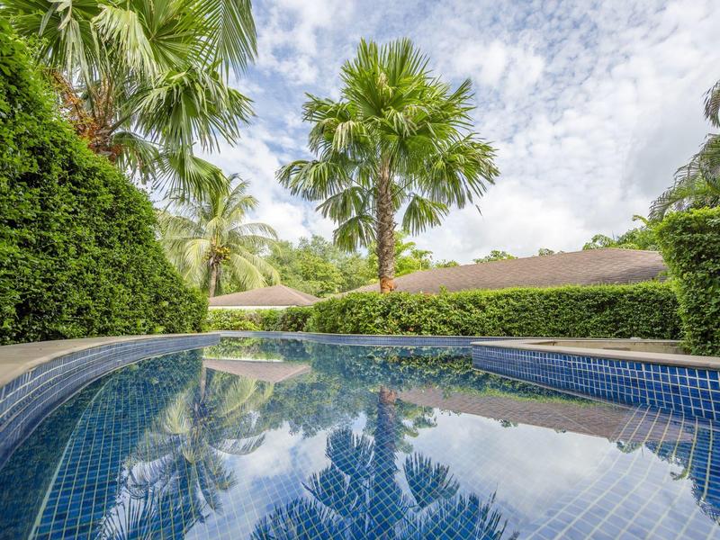 Czysty basen otoczony zielonymi palmami i żywopłotami pod pochmurnym niebem.