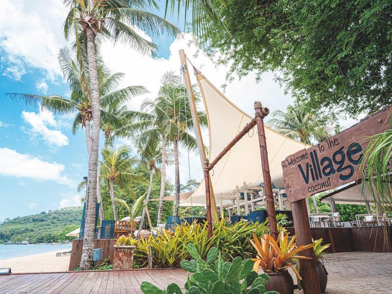 Ośrodek na plaży z drewnianym molo otoczonym palmami, żaglówka i drewniany znak wioski pod błękitnym niebem.