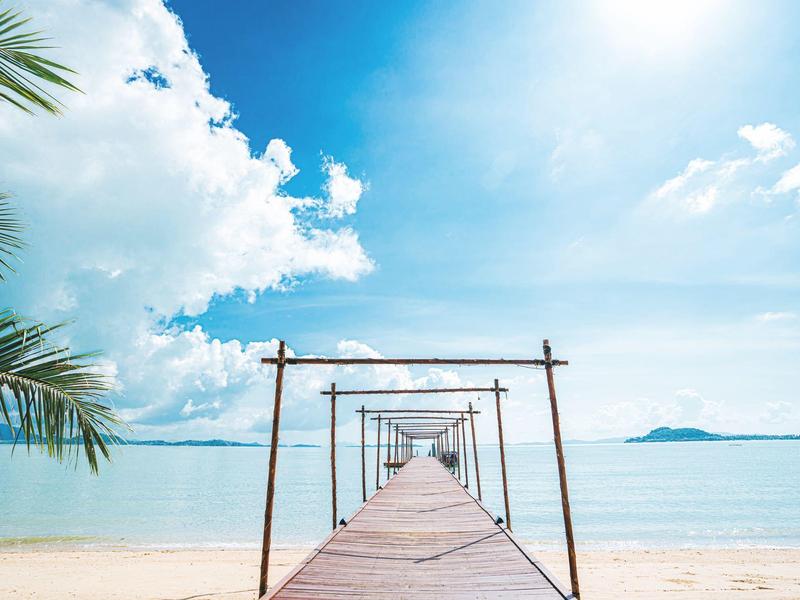 Długi drewniany pomost biegnie przez biały piaszczysty brzeg ku turkusowemu morzu pod niebieskim niebem.