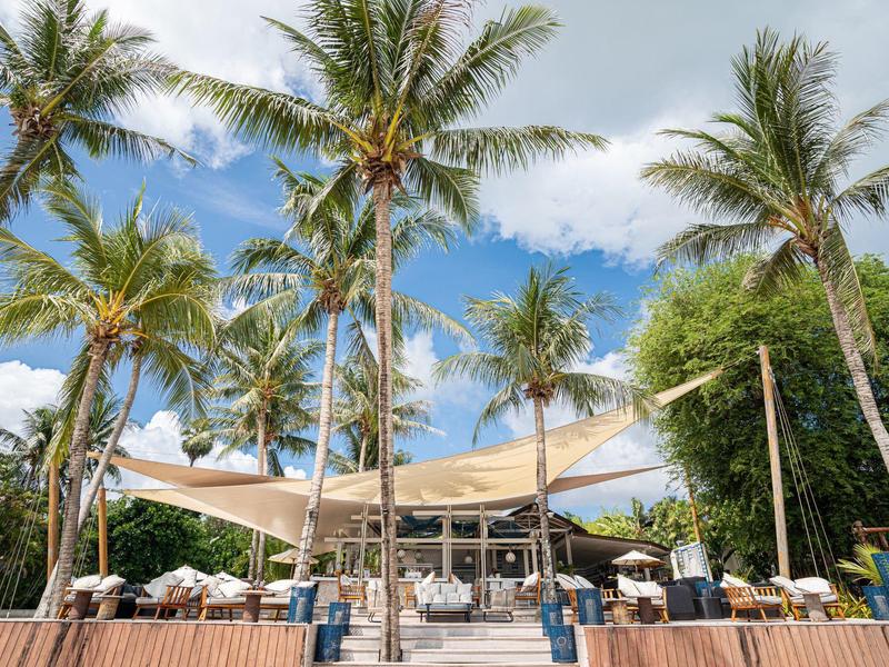 Restauracja przy plaży z żaglami słonecznymi i palmami pod niebieskim niebem.