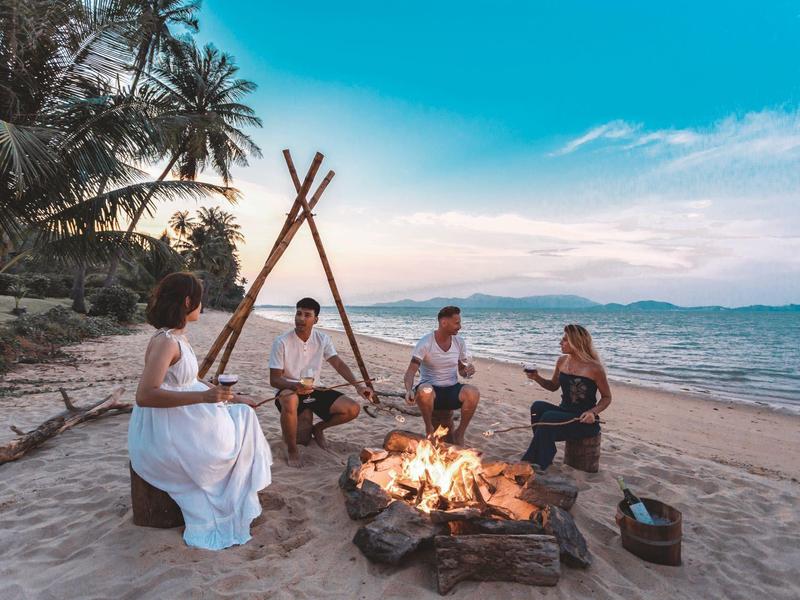 Grupa czterech osób siedzi wokół ogniska na plaży o zachodzie słońca.