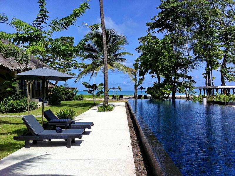 Ligstoelen naast een infinity-zwembad met uitzicht op zee en palmbomen onder een heldere hemel.