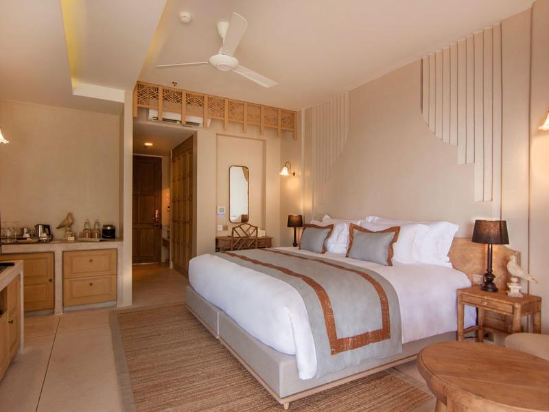 Chambre d'hôtel élégante avec grand lit, tons boisés chauds et ventilateur de plafond moderne.