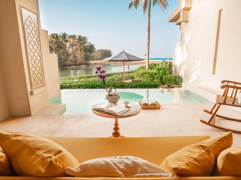 Terrasse confortable avec coussins, vue sur piscine et jardin avec palmiers sous un soleil agréable.