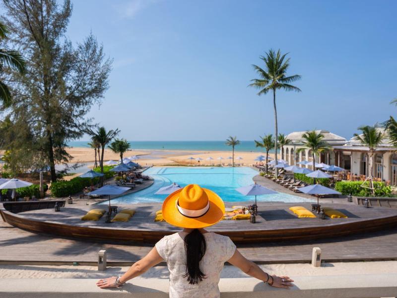 Femme avec chapeau regardant une piscine avec des transats et des palmiers près de la plage.