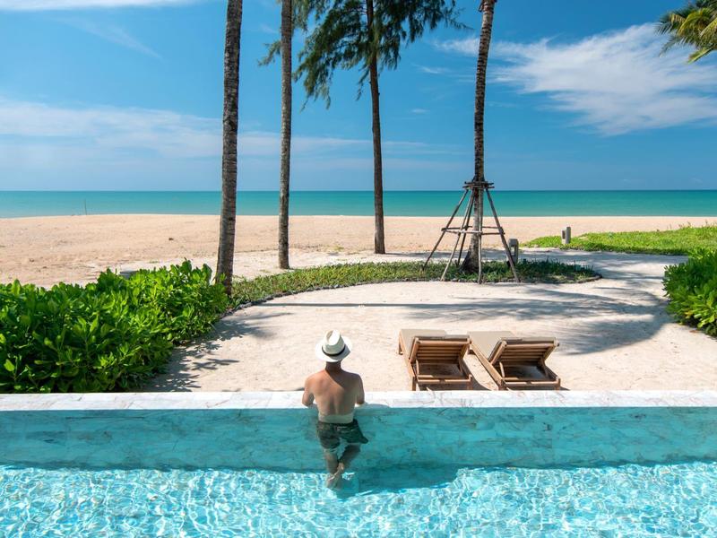 Personne se détendant dans une piscine avec vue sur une plage tropicale de sable et la mer bleue.