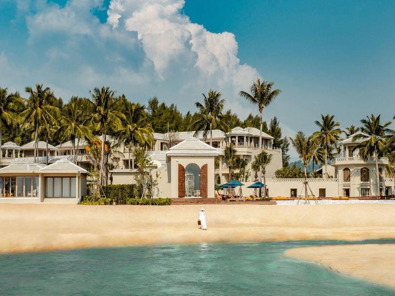Grand hôtel en bord de mer avec des palmiers, sable jaune et mer bleue sous un ciel clair.
