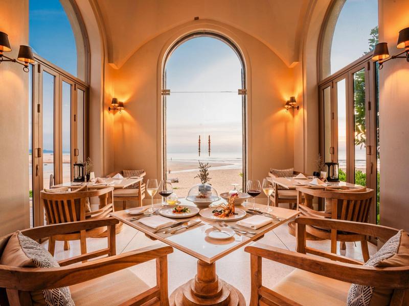 Salle à manger confortable avec mobilier en bois et vue sur la mer à travers de grandes fenêtres cintrées.