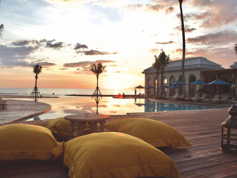 Coucher de soleil au bord d'une piscine avec coussins jaunes sur terrasse en bois et palmiers.