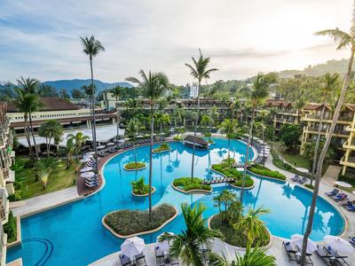 Duży basen hotelowy z wysepkami, palmami i leżakami otoczony kilkoma budynkami oraz górami w tle.