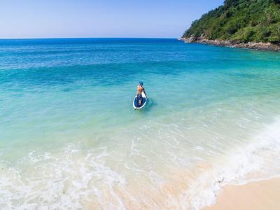 Osoba na desce do paddle w czystym, niebieskim morzu przy piaszczystej plaży i zalesionym brzegu.