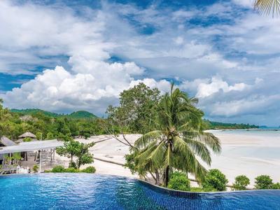 Ośrodek z basenem infinity, palmami i białą piaszczystą plażą pod pochmurnym niebem.