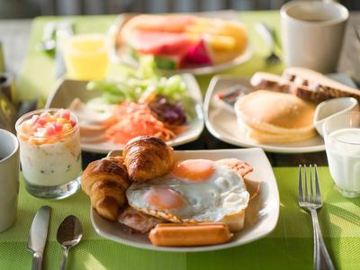 Colazione con uovo fritto, croissant, salsiccia, yogurt e varie bevande su un tavolo verde.