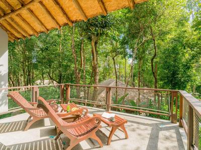 Große Terrasse mit Holzliegen und Tisch, umgeben von einem Wald mit grünem Laub.