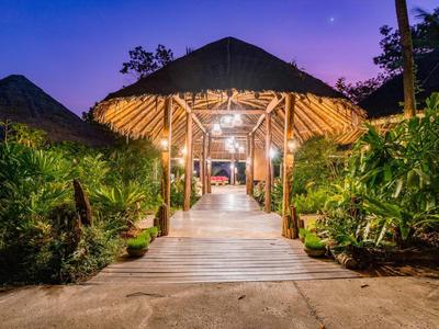 Passaggio in legno illuminato conduce a una capanna aperta con tetto di paglia, circondata da vegetazione rigogliosa.