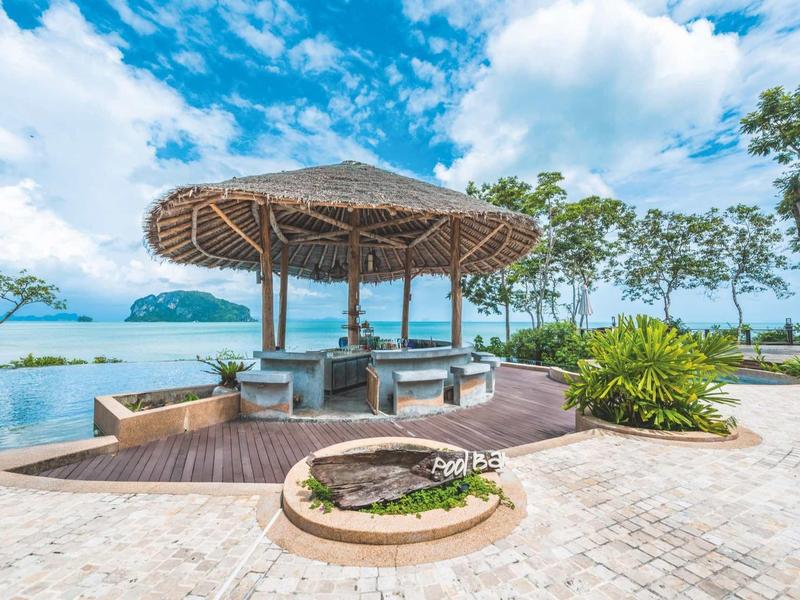 Padiglione aperto con tetto di paglia vicino al mare, circondato da palme e cielo azzurro.