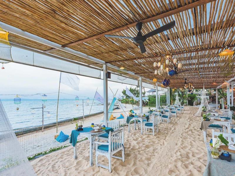 Ristorante aperto sulla spiaggia con tavoli sulla sabbia e vista mare sotto un tetto di legno.