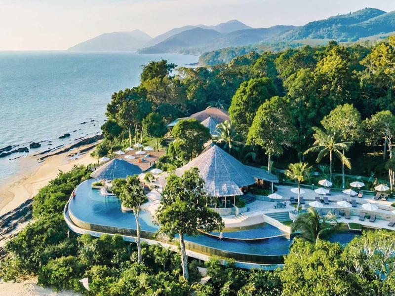 Vista aerea di un resort con piscina e spiaggia, circondato da vegetazione folta e montagne sullo sfondo.