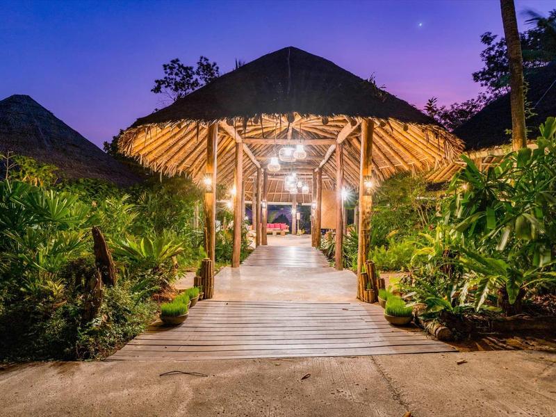 Passaggio in legno illuminato conduce a una capanna aperta con tetto di paglia, circondata da vegetazione rigogliosa.