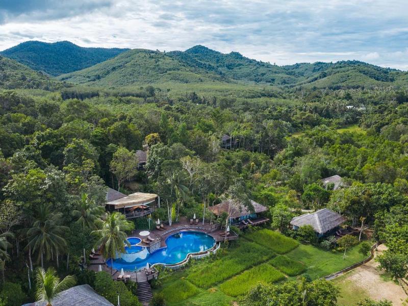 Vista aerea di un hotel con piscina, circondato da una fitta vegetazione e montagne sullo sfondo.