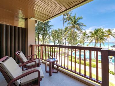 Balcony with chairs and table overlooking pool, garden, and palm trees under a blue sky.