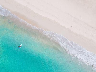 Aerial view of a lone surfer in turquoise water by a white sandy beach.