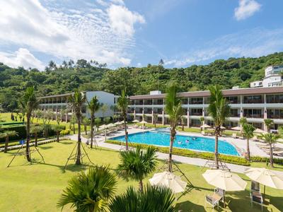 Hotel with clear pool, palm trees, and green hillside backdrop under a blue sky.