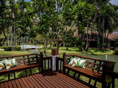 Dřevěná terasa s lavičkami a polštáři, obklopená zelení a rybníkem.