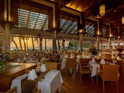 Prostorná, stylová restaurace s dřevěným nábytkem, bílými ubrusy a velkými okny s výhledem na palmy.