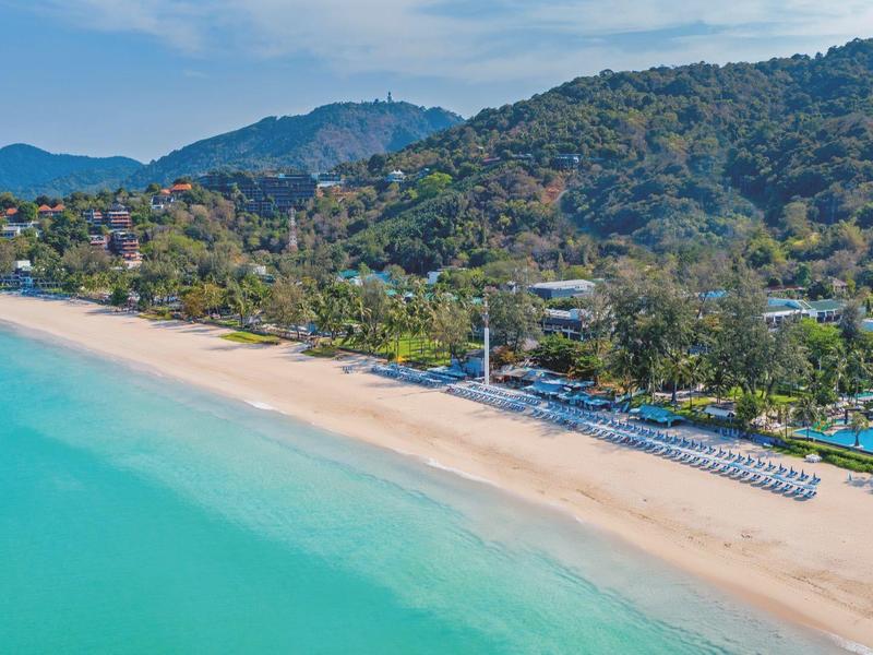 Long, quiet sandy beach beside a hotel with forested hills in the background.