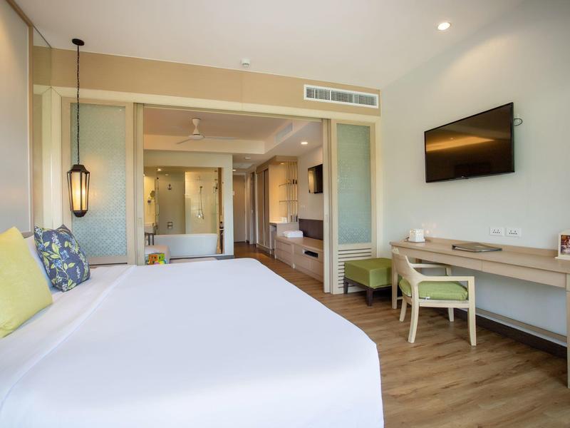 Brightly furnished hotel room with large bed, TV, desk, and bathroom in the background.