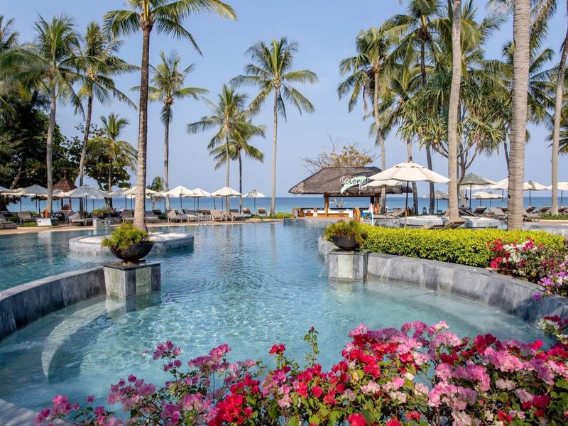 Balcony with chairs and table overlooking pool, garden, and palm trees under a blue sky.
