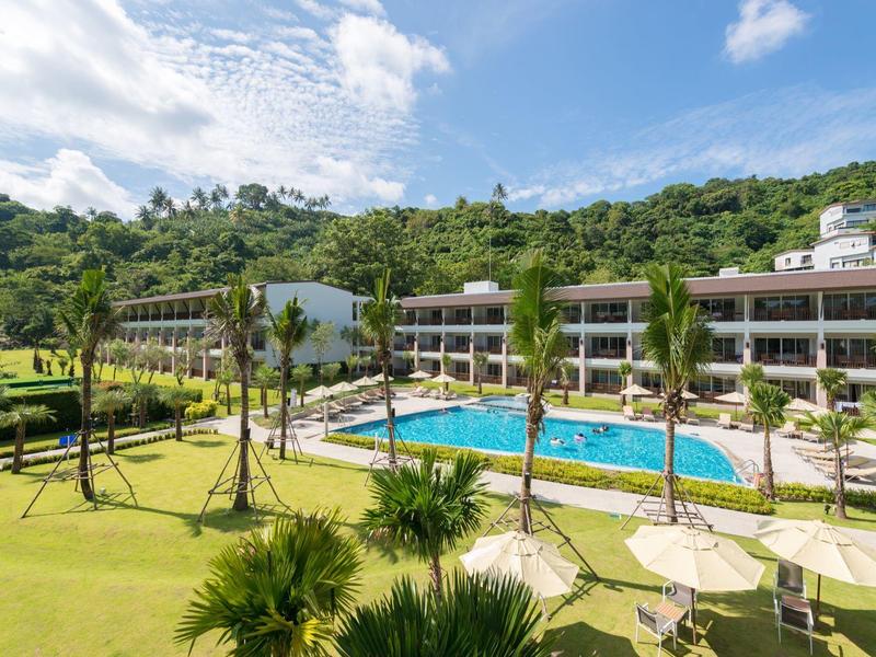 Hotel with clear pool, palm trees, and green hillside backdrop under a blue sky.