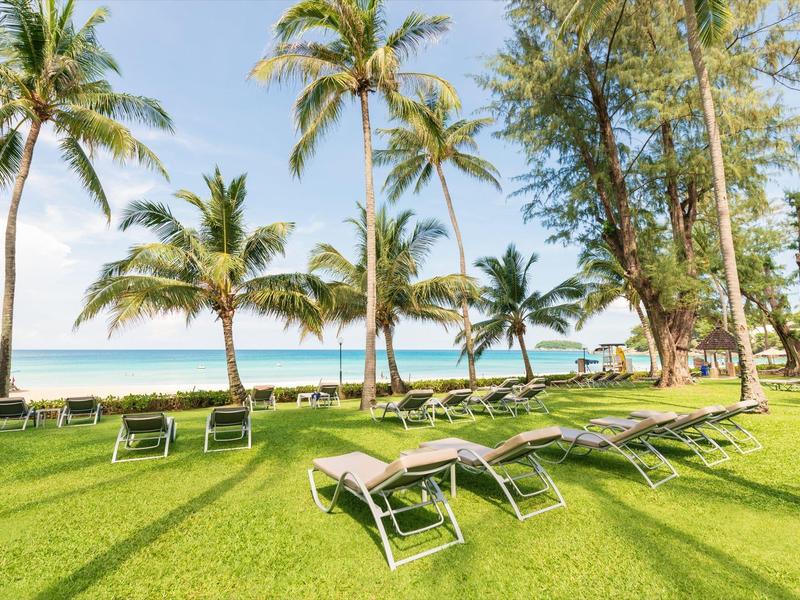 Green lounge chairs on grass with palm trees and sea in the background at a sunny beach.