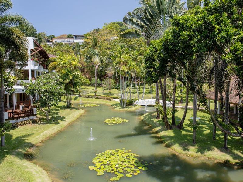 Green hotel garden with pond, fountain, and tropical trees under a blue sky.
