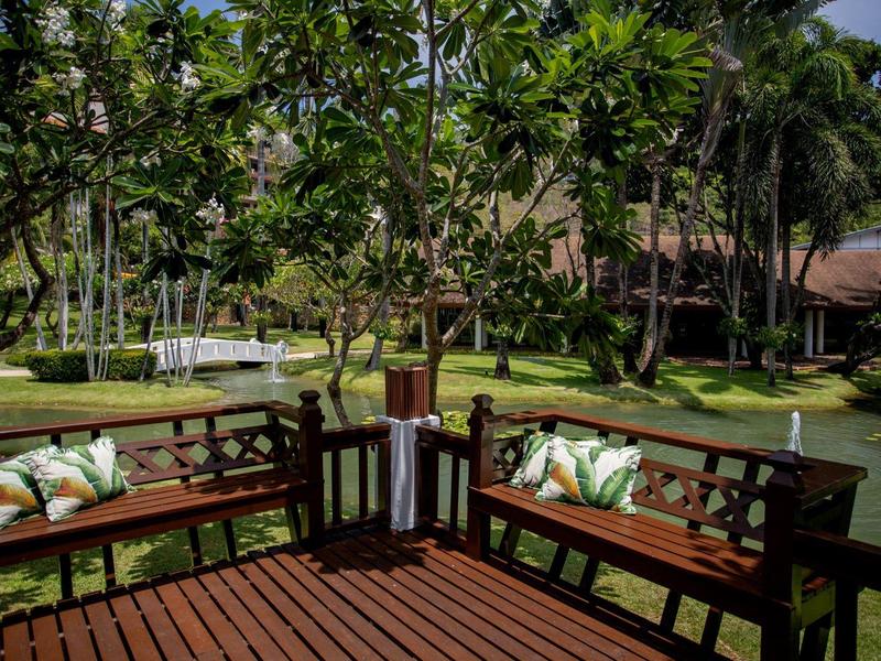 Wooden terrace with benches and cushions surrounded by trees and green landscape in hotel garden.