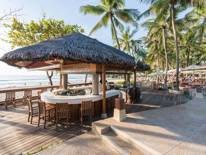 Thatched roof bar on the beach, surrounded by chairs on wooden deck with palm trees in the background.
