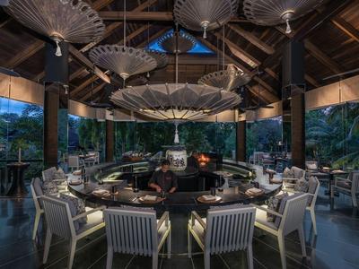 Otwarta jadalnia z nowoczesnym kominkiem i krzesłami pod drewnianym dachem w tropikalnym otoczeniu.