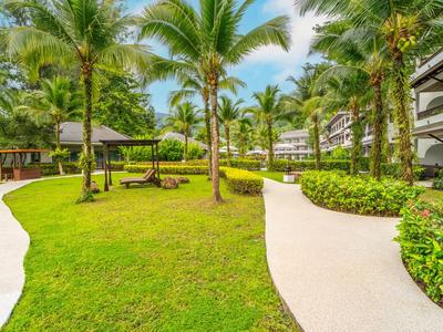 Green garden with palm trees and paved paths between hotel buildings under blue sky.