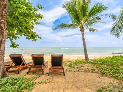 Three lounge chairs on sand under palm trees facing the sea on a clear day.