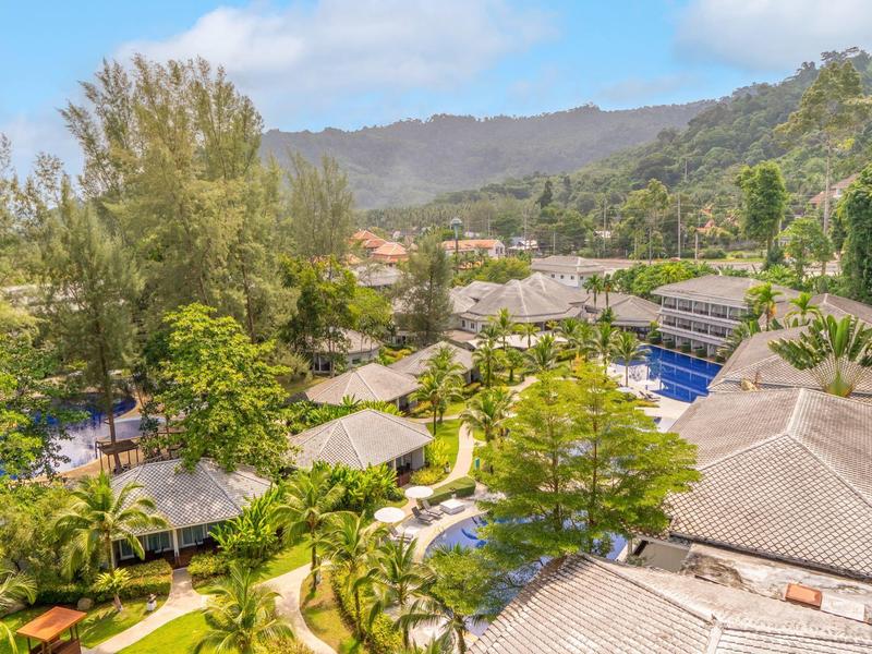 Hotelcomplex met zwembaden, groene paden en hutten tegen een bergachtergrond bij blauwe lucht.