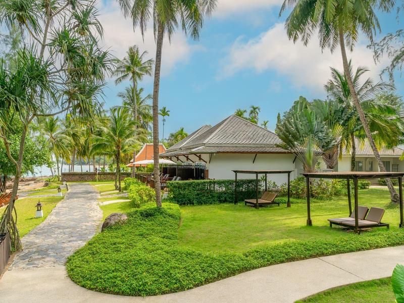 Bungalow omgeven door palmbomen en gras in tropisch resort onder zonnige lucht.