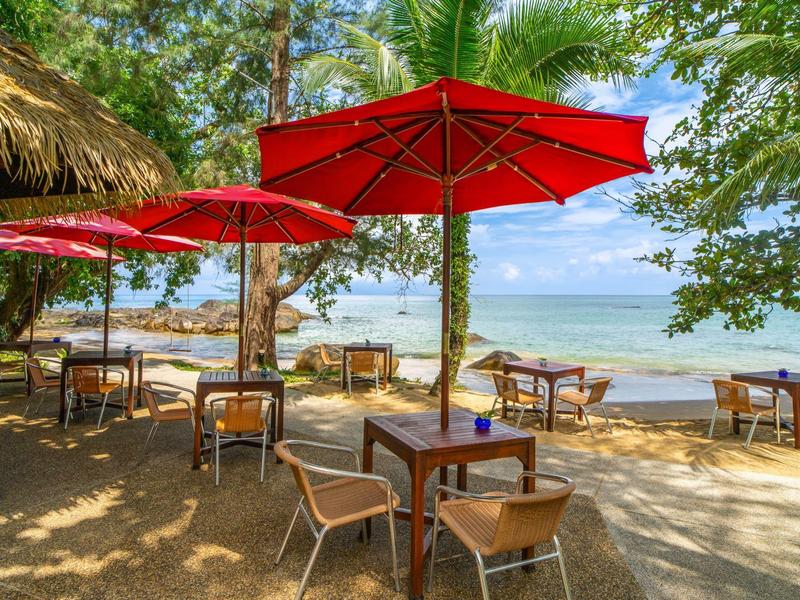 Strandrestaurant met tafels en rode parasols, uitzicht op zee onder blauwe lucht.