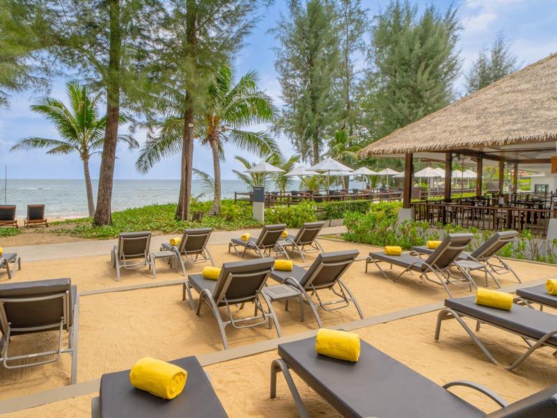 Ligstoelen met gele handdoeken op het zandstrand, palmbomen en een paviljoen op de achtergrond.