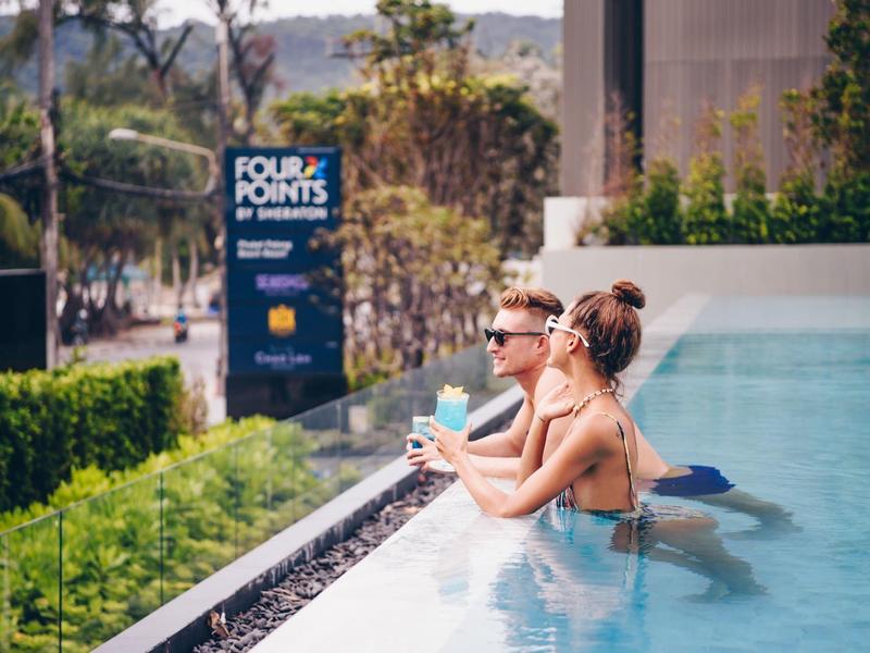 Dwie kobiety cieszą się koktajlami przy hotelowym basenie otoczonym zielenią.