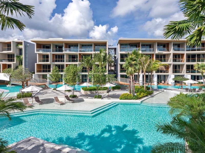 Complesso alberghiero moderno con piscina, ombrelloni e palme sotto un cielo azzurro.