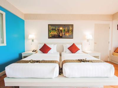 Licht ingericht tweepersoonskamer met witte bedden, rode kussens en een kleurrijk schilderij aan de muur.