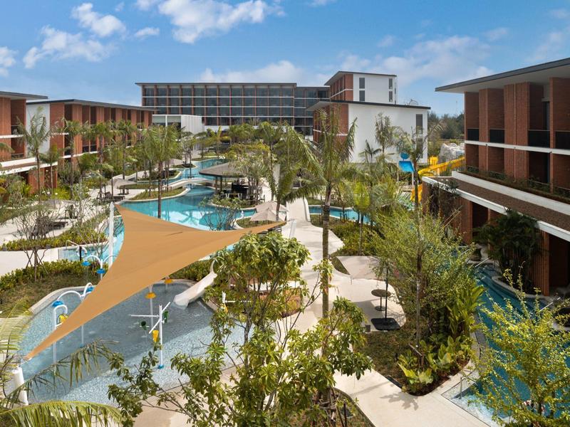 Complejo hotelero moderno con piscinas, tumbonas y vegetación tropical bajo un cielo azul.