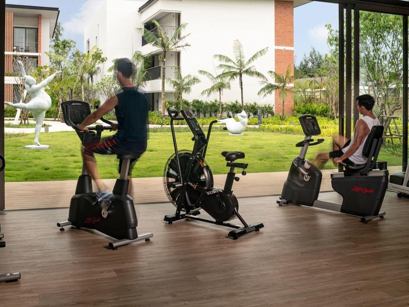 Personas entrenan en bicicletas estáticas en un gimnasio con vista a un jardín.